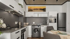 Une cuisine moderne équipée d’un lave-linge intégré, un coin repas avec une table pour deux personnes, et un grand tableau accroché au-dessus de l’évier.