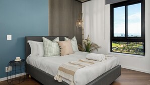 Une chambre moderne avec un grand lit, une vue sur le paysage urbain par la fenêtre et une petite table d’appoint sur laquelle se trouve un bol.