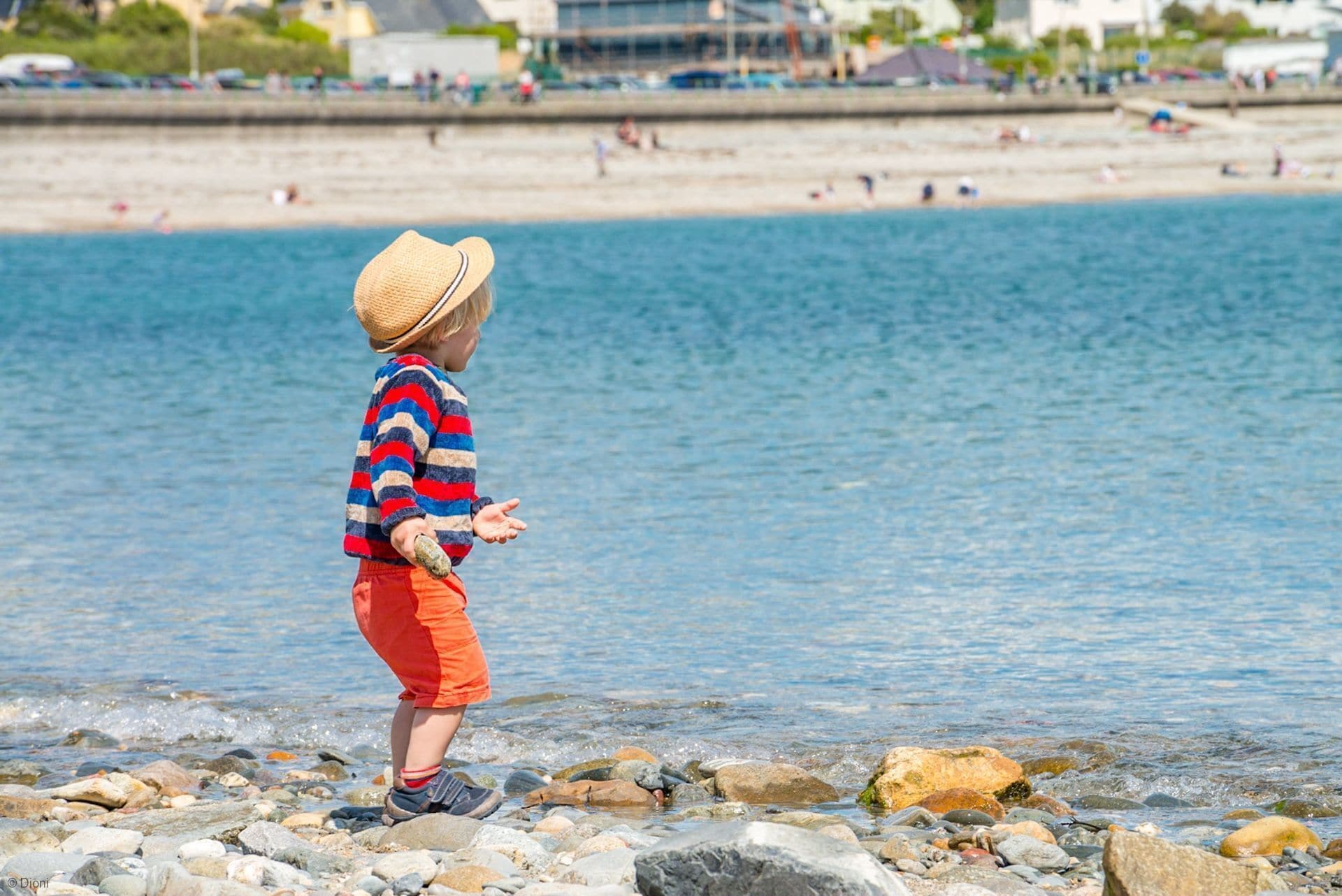 A child playing by a rocky beach with a clear view of the sea and distant buildings.