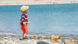 A child playing by a rocky beach with a clear view of the sea and distant buildings. - Luxurious Three Bedroom Farmhouse with Sea Views in Llanystumdwy | Bryn-Du (Criccieth)