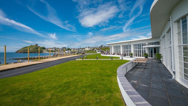 A modern building with large windows, a grassy lawn, and a view of the sea and a hill with a castle. - Luxurious Three Bedroom Farmhouse with Sea Views in Llanystumdwy | Bryn-Du (Criccieth)