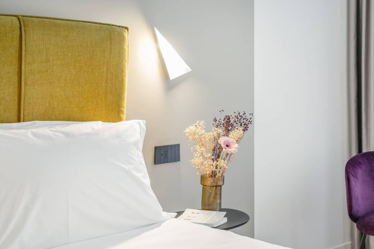 Quarto de hotel com cama, mesa de cabeceira com um vaso de flores, abajur e uma cadeira roxa.