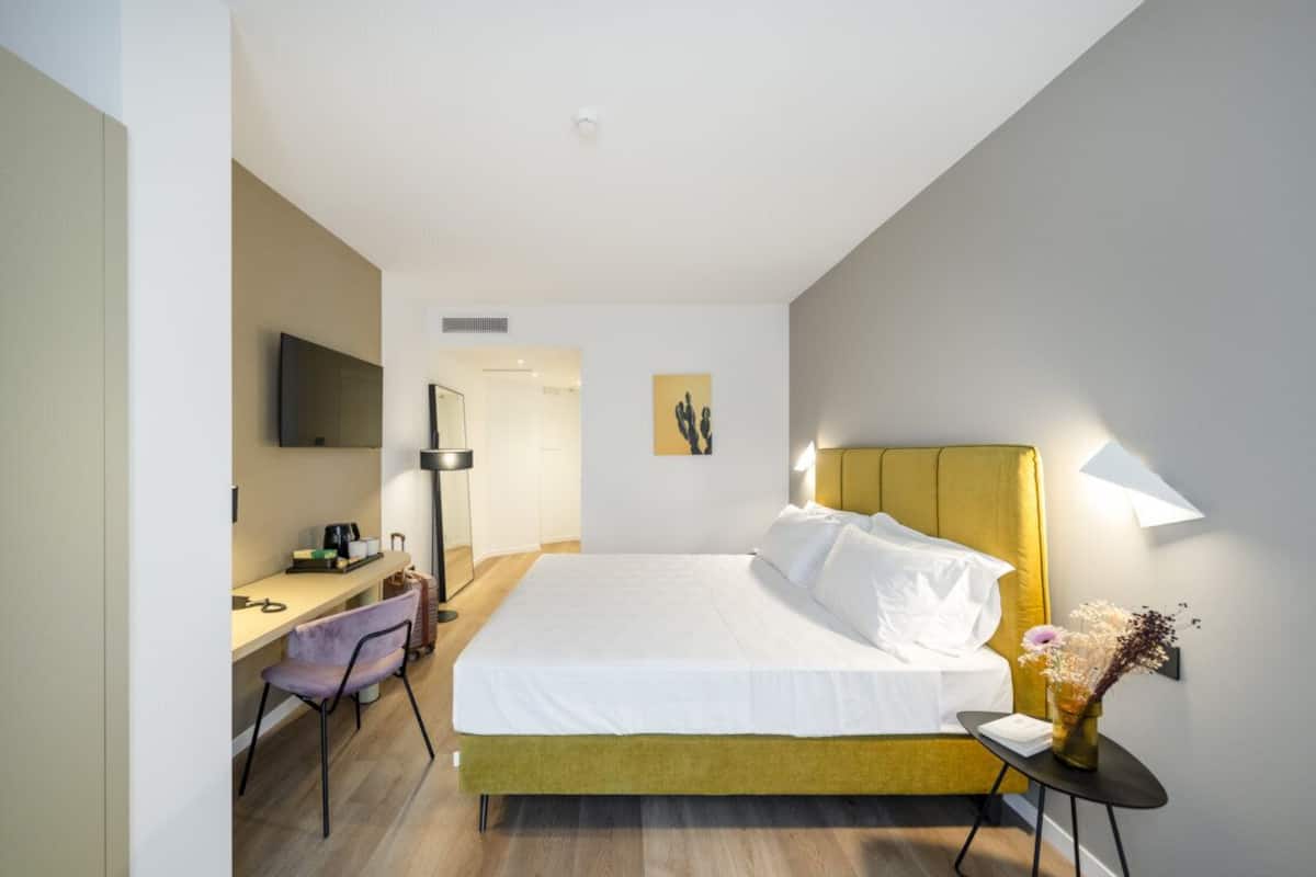 Quarto de hotel com cama, escrivaninha, cadeira, televisão e luminária na parede.