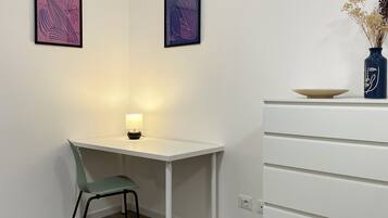A small, white desk with a green chair, a vase with dried flowers, and two framed artworks on the wall.