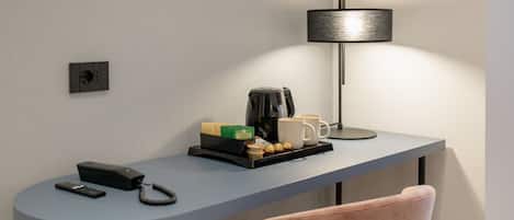A modern hotel room with a desk, a lamp, a coffee maker, and a chair.