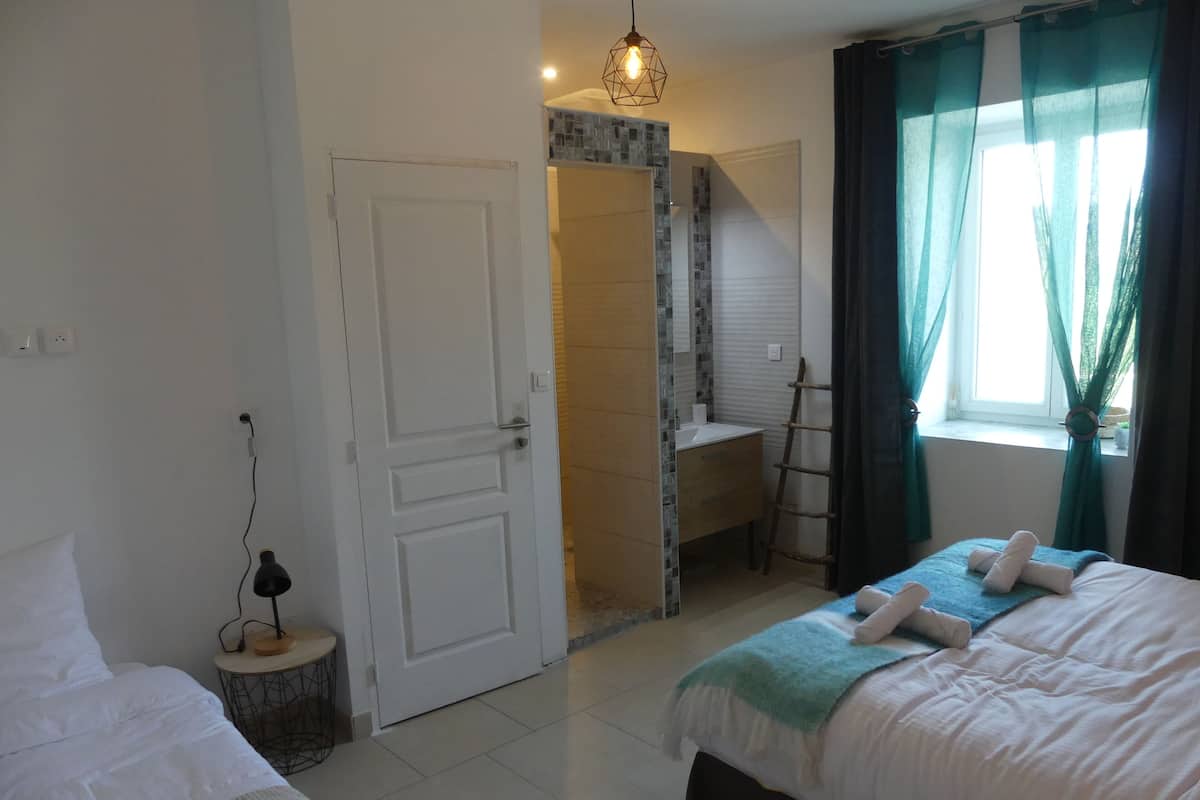A hotel room with a bed, a bedside table with a lamp, a window with curtains, and a bathroom visible through an open door.