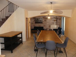 Un comedor con una larga mesa de madera y seis sillas, una escalera a la izquierda, y una pared de ladrillo con un cuadro en el fondo.
