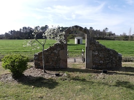Un arco de piedra que da acceso a un pequeño edificio blanco con una puerta oscura, rodeado por un campo cubierto de hierba y árboles.