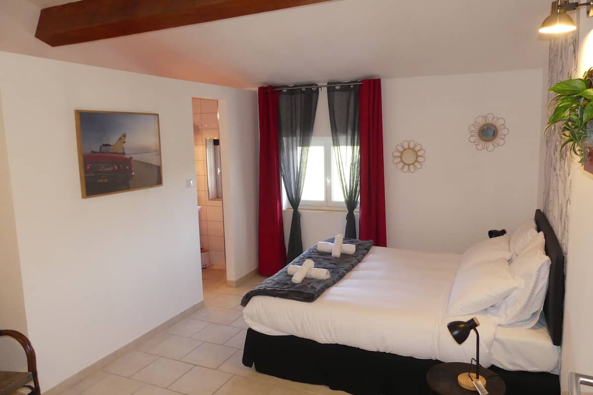 A bedroom with a bed, a chair, a bedside table with a lamp, a painting on the wall, and a window with curtains.