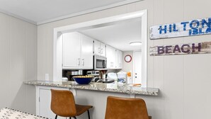 A kitchen with white cabinets, a microwave, and a bowl on the counter. There are two brown chairs and a patterned couch. A sign on the wall reads 'Hilton Island Beach.' - Gorgeous Oceanfront Villa, w/ Pool on Hilton Head (Hilton Head Island)