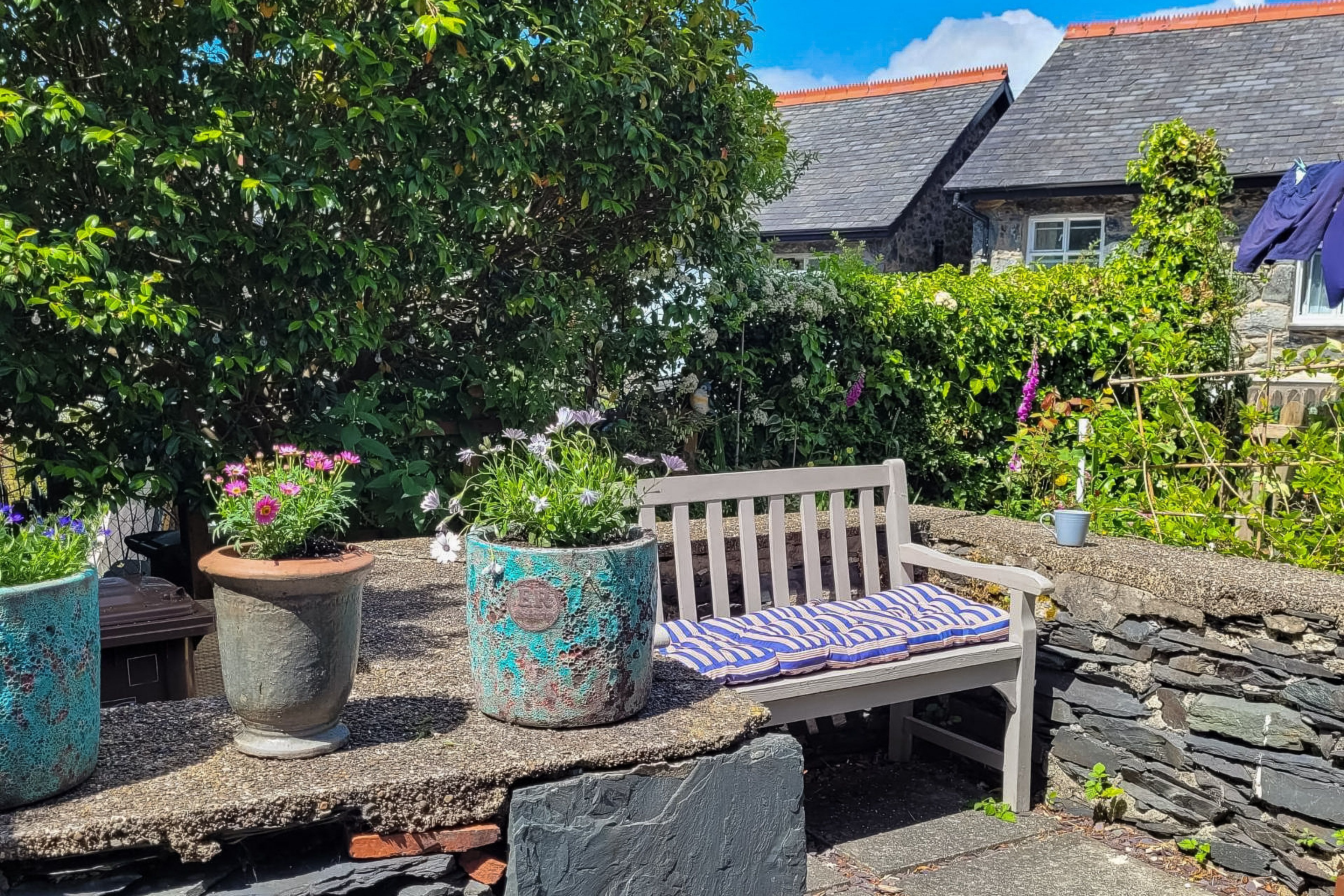 A stone bench with a patterned cushion in a garden area with potted plants and a stone wall.