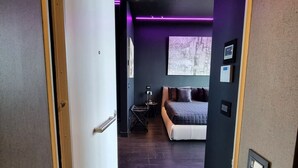 Ein modernes Hotelzimmer mit dunklem Holzboden, einem Bett mit grauer Kopfteil und einem kleinen Schreibtisch mit Stuhl.