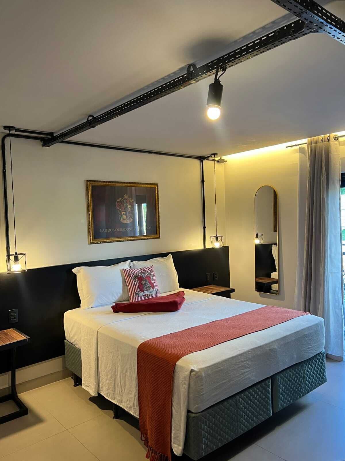 Quarto de hotel com uma cama, mesa de cabeceira, espelho e um quadro na parede.