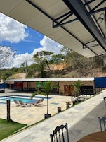 Uma área externa coberta com piscina, espreguiçadeiras e um alpendre com mesas e cadeiras.