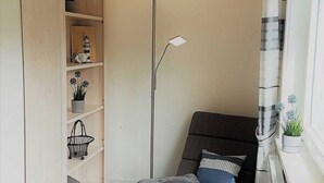 A corner room with a grey sofa, a floor lamp, a window with curtains, and a shelf with decorative items. - Haus Mira - Wohnung Süden (Wittdün auf Amrum)
