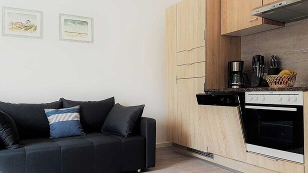 A modern kitchen with wooden cabinets, a black dishwasher, and a stove. There is a black sofa with cushions and two framed pictures on the wall. - Haus Mira - Wohnung Süden (Wittdün auf Amrum)