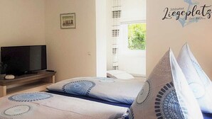 A bedroom with two beds, a television on a stand, and a window with curtains. - Haus Mira - Wohnung Süden (Wittdün auf Amrum)