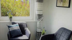 A room with two grey upholstered chairs, a small table, and a window with a view of greenery. - Haus Mira - Wohnung Süden (Wittdün auf Amrum)