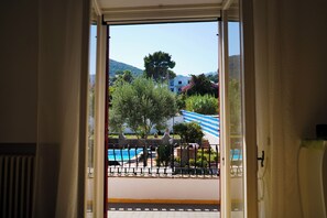 An open door to a balcony with a view of a swimming pool, greenery, and buildings in the background.
