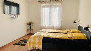 Een slaapkamer met een bed, een televisie, een klein tafeltje met een vaas en een raam met gordijnen.