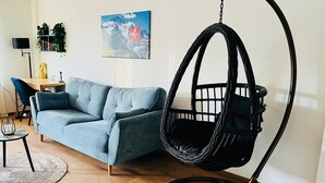 Een woonkamer met een blauwe bank, een hangstoel, een houten vloer, een schilderij aan de muur, en een logo onderaan.