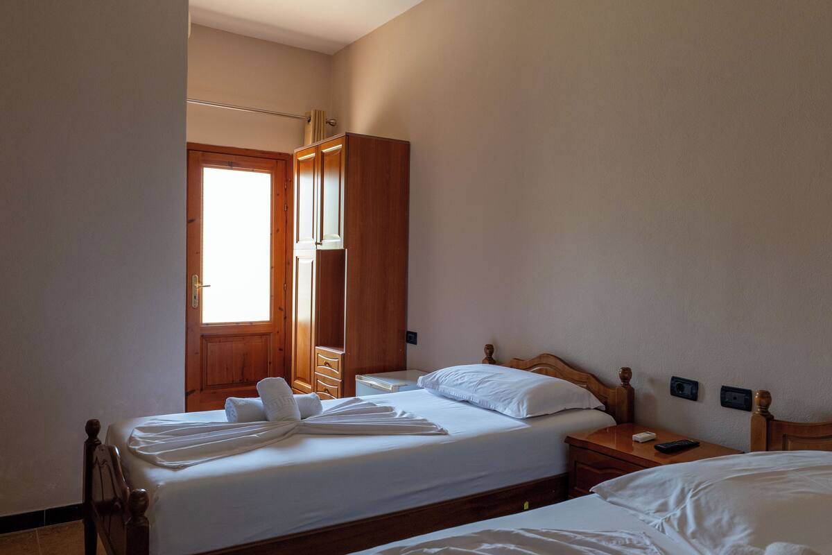 A hotel room with two beds, wooden headboards, a wooden wardrobe, a bedside table with a lamp, and a window with natural light.