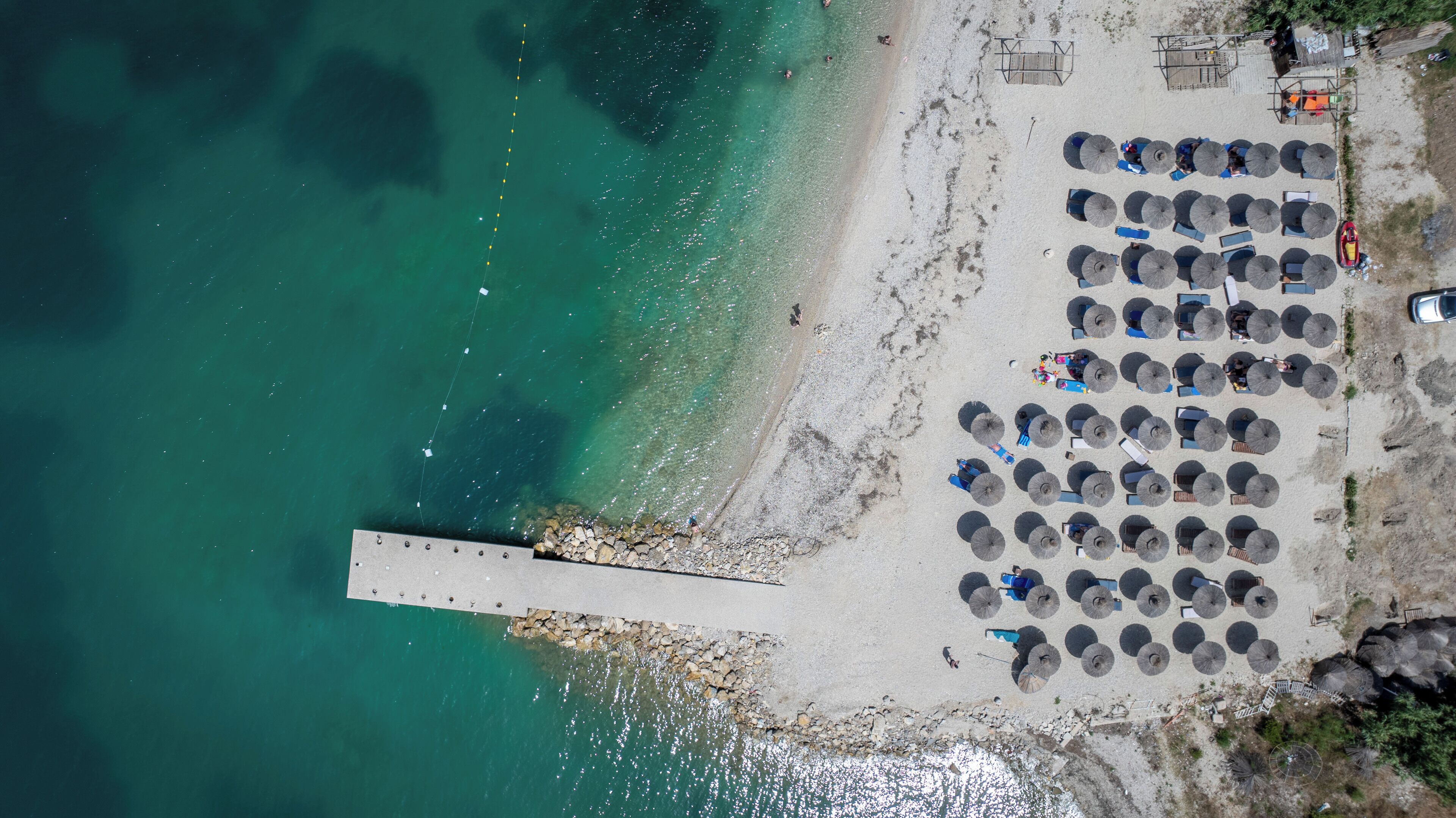 An aerial view of a beach with a pier extending into the water, rows of umbrellas, and a few people.