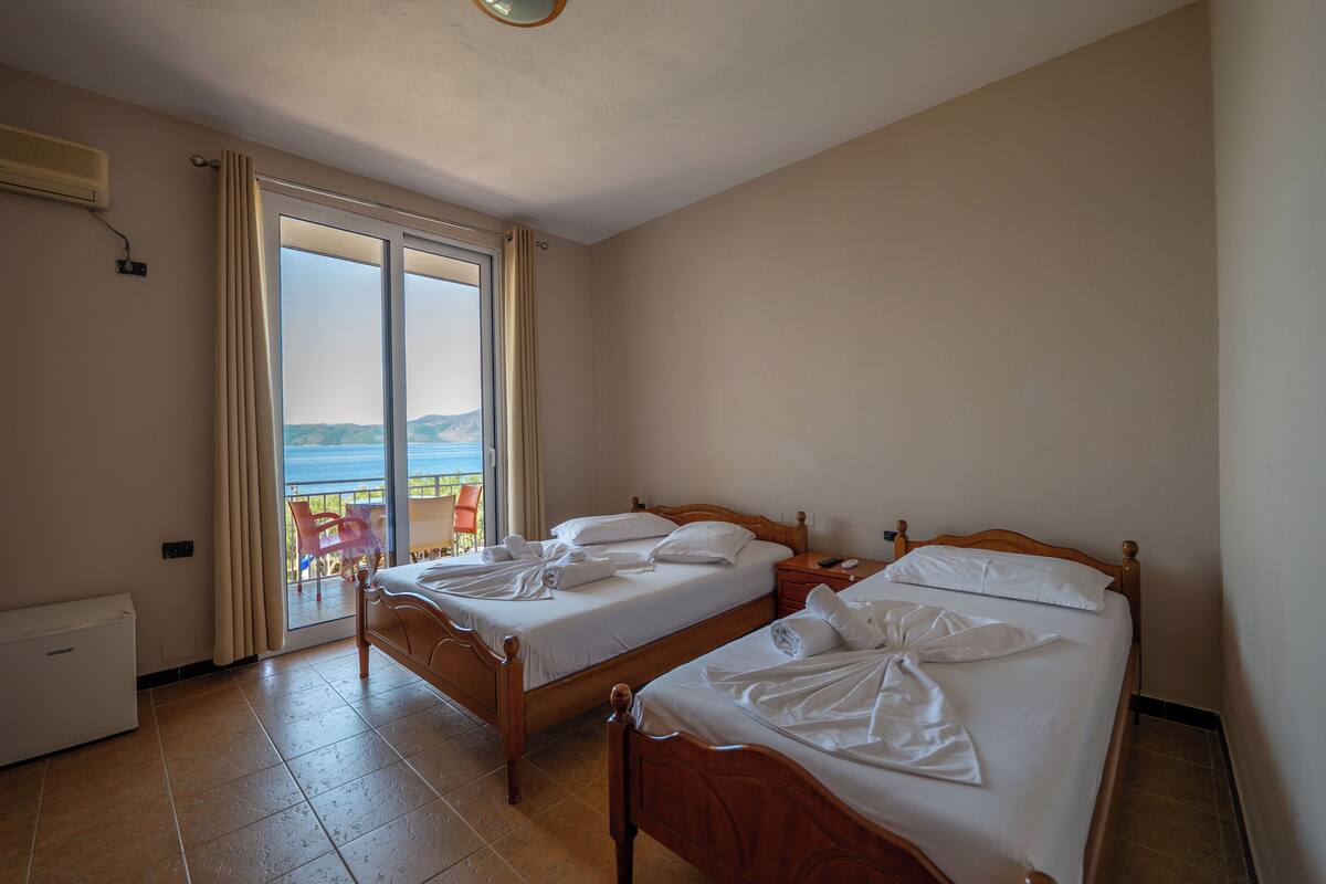 A hotel room with two wooden beds, a sliding glass door to a balcony, and a built-in air conditioner.