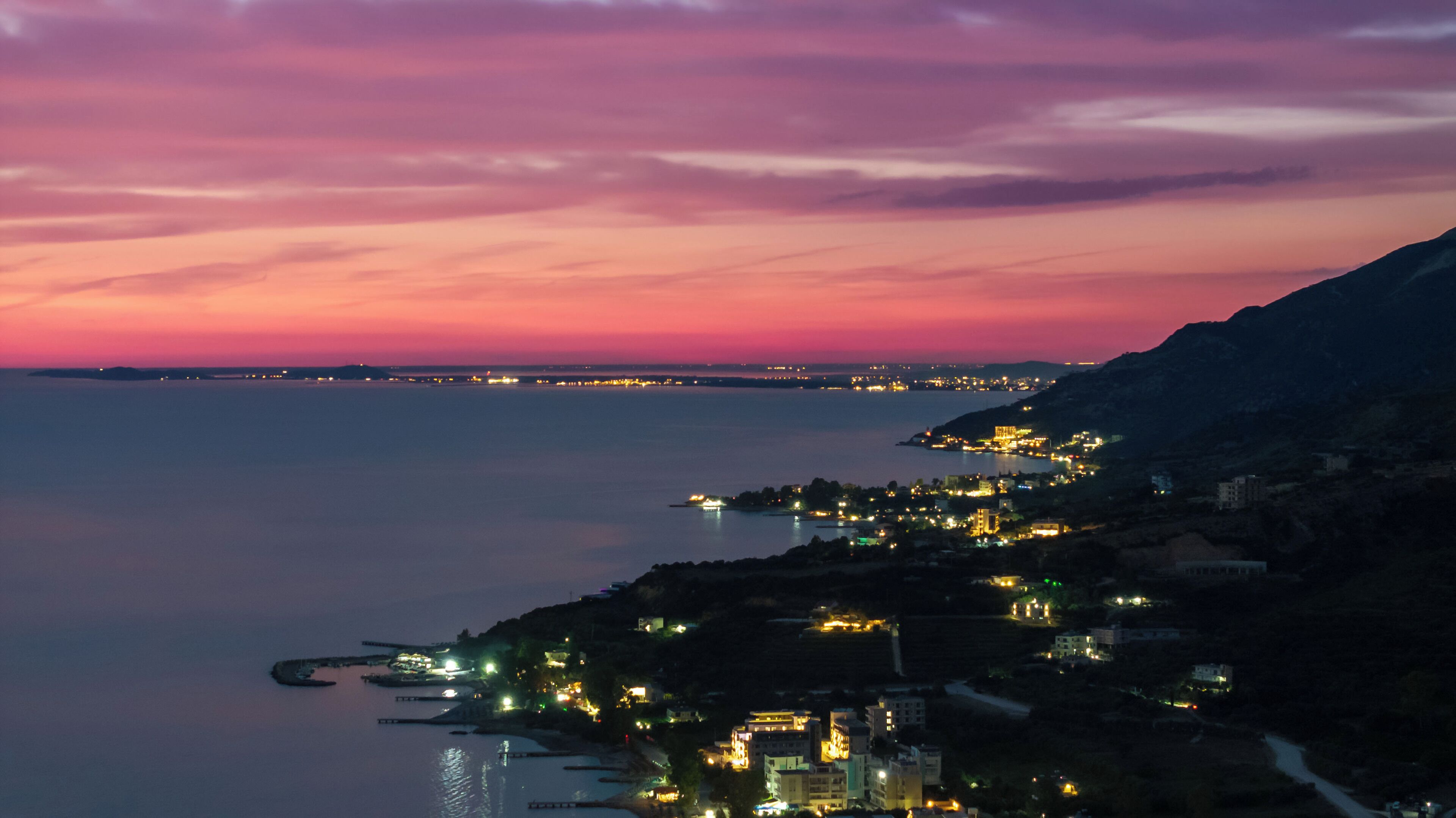 A coastal town at dusk with buildings along the shoreline and a mountain in the background.