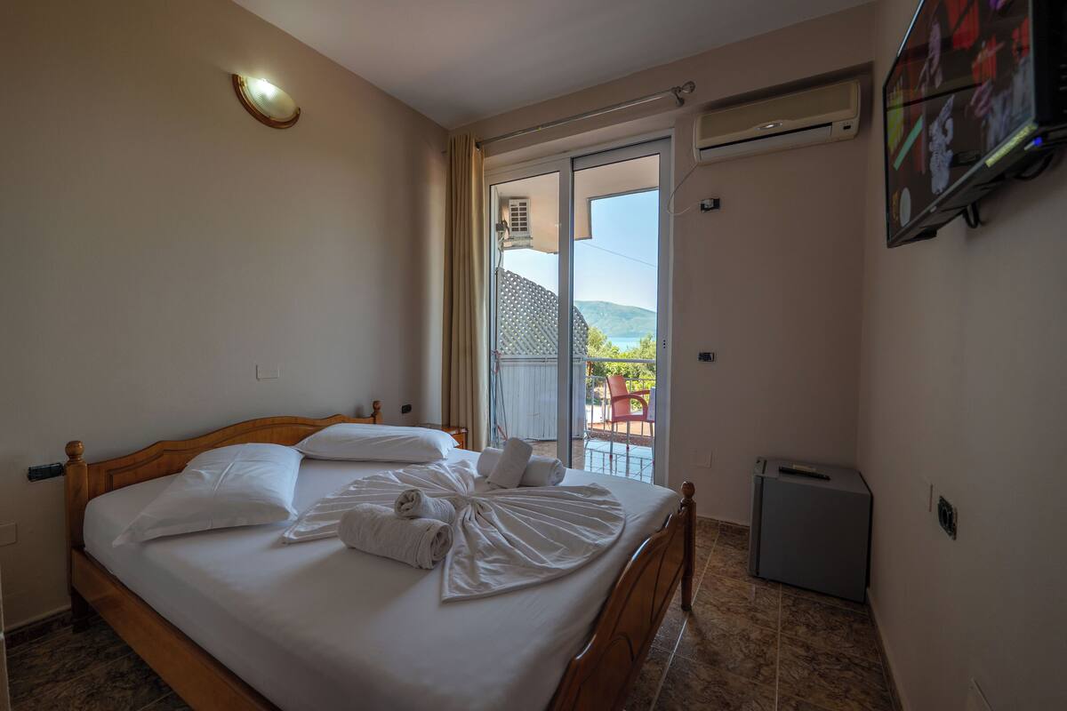 A hotel room with a bed, a TV, a small trash bin, and a sliding glass door to the outside.