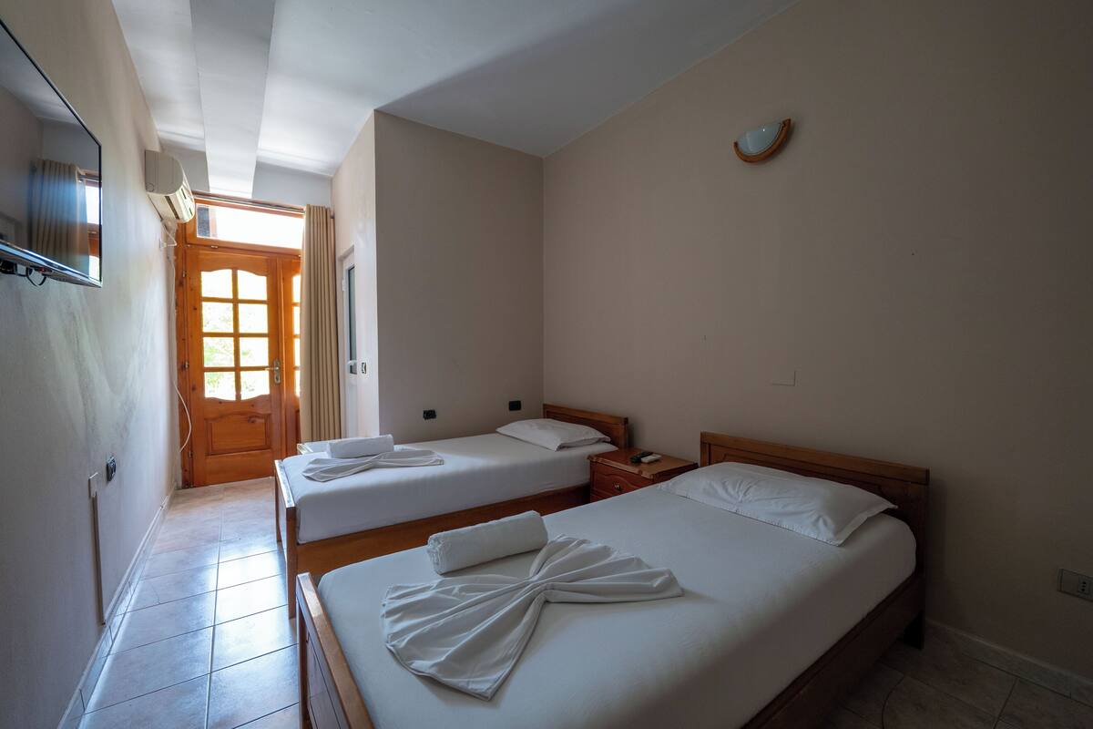 A hotel room with two beds, a wooden headboard, a bedside table, a nightstand, a mirror, and a window with curtains.