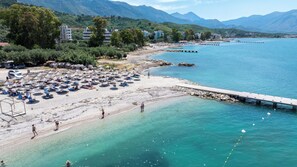 Una playa con tumbonas y sombrillas, un muelle que se adentra en el agua, y edificios al fondo.