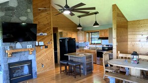 A rustic cabin interior with a stone fireplace, wooden ceiling beams, a kitchen with a refrigerator, stove, and wooden cabinets, and a dining area with benches. - Breathtaking Views!  Spacious Cabin, HotTub, pool table near PF & Gatlinburg ⛰️ (Sevierville)
