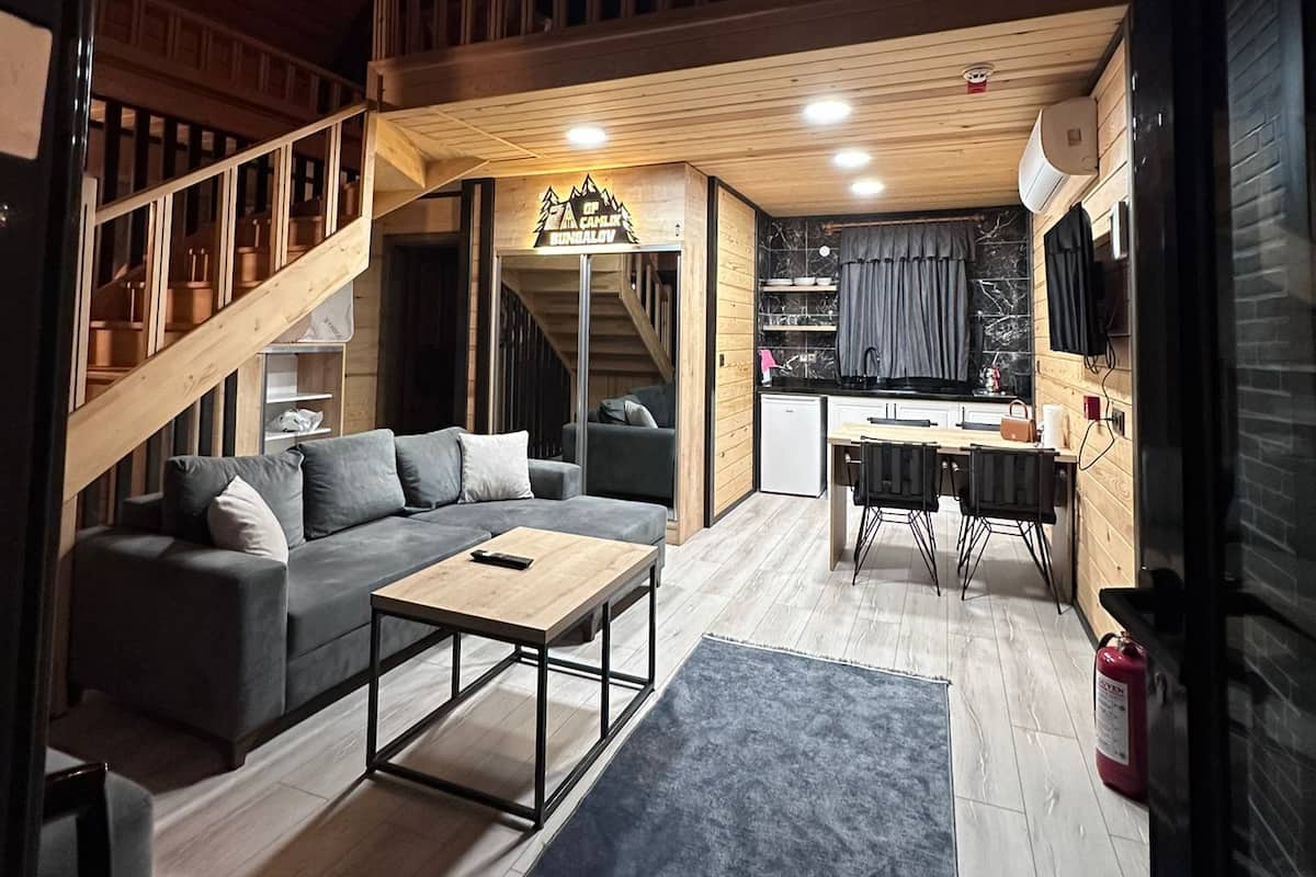 Un área de estar amplia con techo alto, vigas de madera, una escalera, un sofá, una mesa de centro, una cocina y una mesa de comedor.
