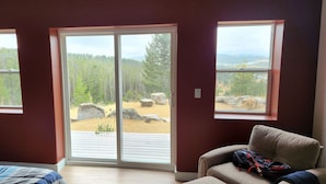 Una habitación con cama, sofá y un amplio ventanal corredero que da a un espacio exterior con terraza de madera y vistas a un bosque.