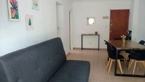 A living room with a dark grey sofa, a wooden dining table with chairs, a small side table, and a door.