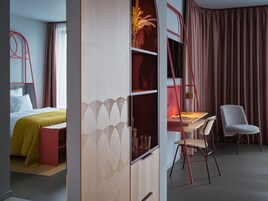Una habitación de hotel moderna con una cama, un escritorio con silla, una estantería y un ventanal con cortinas.