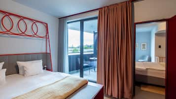 Une chambre d’hôtel moderne avec un grand lit, une tête de lit rouge, un balcon avec vue et une chambre séparée comprenant une chaise et un berceau.