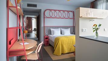 Habitación de hotel pequeña con cama, escritorio y silla. Hay una mesita con lámpara, y una ventana con cortinas.