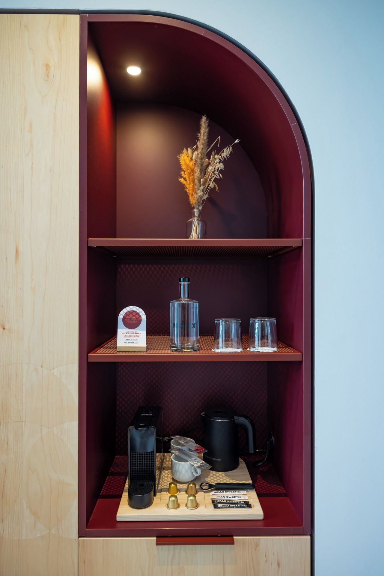 Un mueble de madera con repisa curva que contiene un florero con flores secas, una botella y dos vasos. Abajo, hay dos cafeteras, una tetera y un juego de tres campanas.