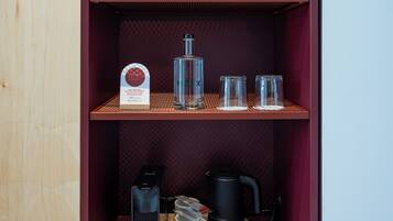 Un mueble de madera con repisa curva que contiene un florero con flores secas, una botella y dos vasos. Abajo, hay dos cafeteras, una tetera y un juego de tres campanas.