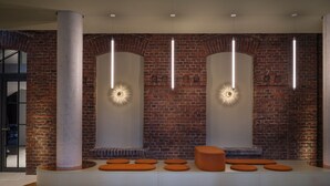 A modern lobby with a brick wall, recessed lighting, and a seating area with orange cushions. - Noxx Hotel (Marburg)