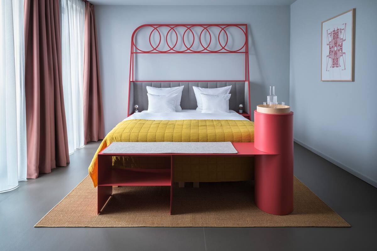 Ein modernes Hotelzimmer mit einem Bett, einem Nachttisch, einem roten, zylindrischen Objekt und einem gerahmten Bild an der Wand.