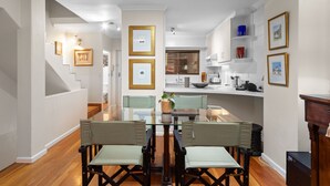 A dining area with a glass table and chairs, a kitchen in the background, and framed pictures on the walls.