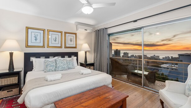A modern bedroom with a large bed, a ceiling fan, and a city view through floor-to-ceiling windows.