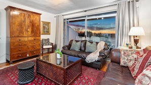 A living room with a brown leather sofa, a wooden cabinet, a glass coffee table, and a view of a sunset through large windows.