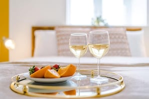 A bed with a white and gold bedspread, two glasses of white wine, and a plate of fruit on a tray.