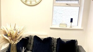 A living room with a grey sofa, a round gold clock, a vase with dried plants, and a window with a view of a brick wall.