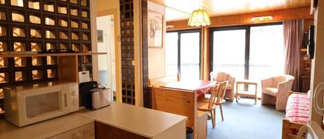A compact kitchen with a microwave, a small refrigerator, and a wooden dining table with chairs. There is a balcony with a view and a hanging light fixture.