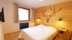 A bedroom with a large bed, a window with curtains, a radiator, a wall-mounted light fixture, and a wooden wall with a tree-like design. - Wifi, 200m from ski slopes, 1st floor, balcony, 65m², Les Gets (Les Gets)